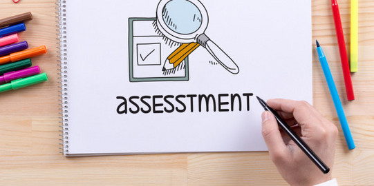 Sheet on desk with drawing and lettering "Assessement", above hand with pencil