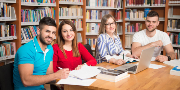 4 Studierende sitzen in einer Bibliothek am Tisch, Blick in die Kamera