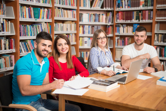4 Studierende sitzen in einer Bibliothek am Tisch, Blick in die Kamera