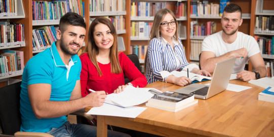 Schreibgruppe 4 Studierende sitzen in einer Bibliothek am Tisch, Blick in die Kamera