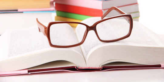 Stapel von Büchern im Hintergrund, aufgeschlagenes Buch und Lesebrille im Vordergrund