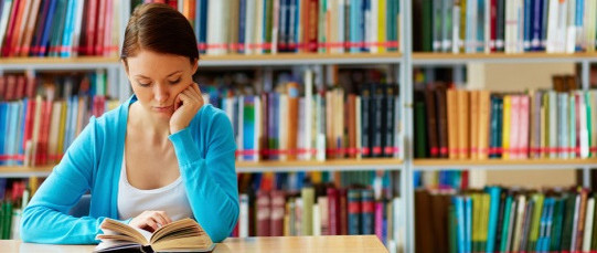Junge Frau lesend in Bibliothek über Buch gebeugt, im Hintergrund Bücherwand