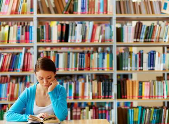 Junge Frau lesend in Bibliothek über Buch gebeugt, im Hintergrund Bücherwand