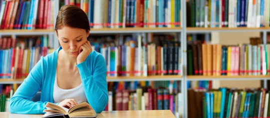 Junge Frau lesend in Bibliothek über Buch gebeugt, im Hintergrund Bücherwand